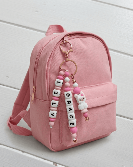 Handcrafted, personalized charm made from BPA-free soft silicone in shades of pink and white. It features three strands: two with name beads and one with a cute white llama figurine with a pink rose, all attached to an elegant rose gold clasp.