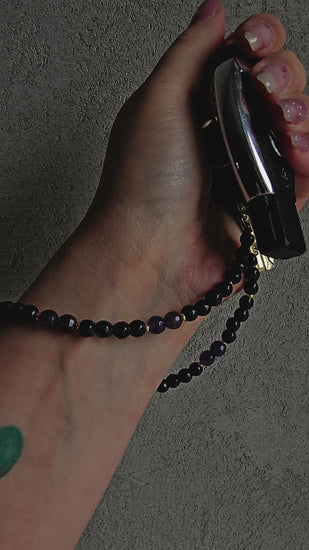 Handmade crystal phone charm with black onyx and amethyst beads, gold accents, and a cord – used as a protective and calming accessory.